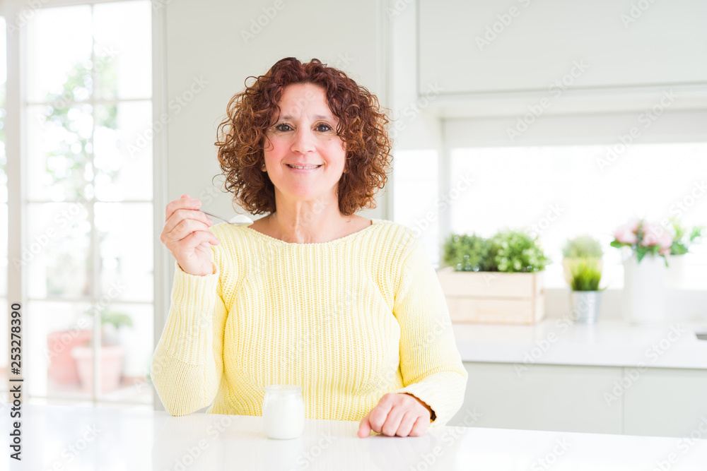 Senior woman eating a healthy natural yogurt at home with a happy face standing and smiling with a confident smile showing teeth