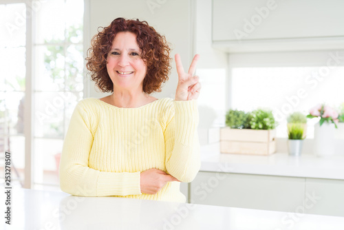 Beautiful senior woman wearing yellow sweater smiling with happy face winking at the camera doing victory sign. Number two.