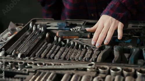 Gentle female hands of a young girl, an auto mechanic, with a manicure and painted nails, against the background of a set of open-end wrenches and other devices for car repair. Girl chooses a wrench