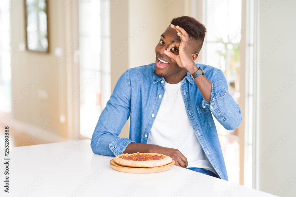 African american man eating pepperoni pizza at home with happy face smiling doing ok sign with hand on eye looking through fingers African american man eating pepperoni pizza at home with happy face smiling doing ok sign with hand on eye looking through fingers