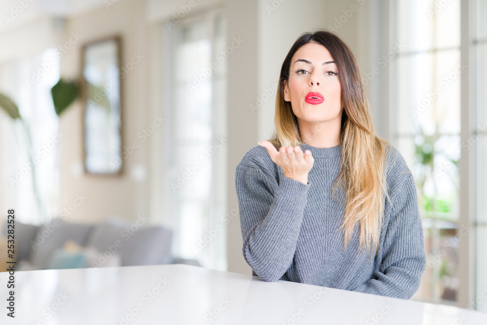 Young beautiful woman wearing winter sweater at home looking at the camera blowing a kiss with hand on air being lovely and sexy. Love expression. Young beautiful woman wearing winter sweater at home looking at the camera blowing a kiss with hand on air being lovely and sexy. Love expression.