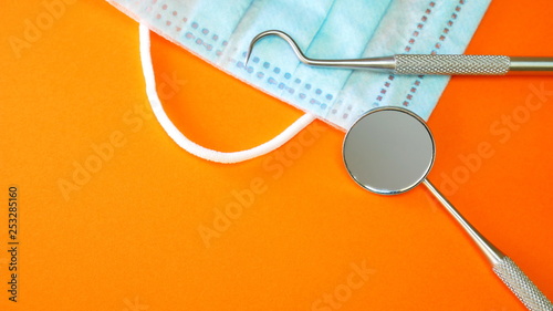 Dentist tools or instruments in dental office: dental explorer, dental mirror and surgical or procedure face mask on orange surface for copyspace text. Dental hygiene and health conceptual image