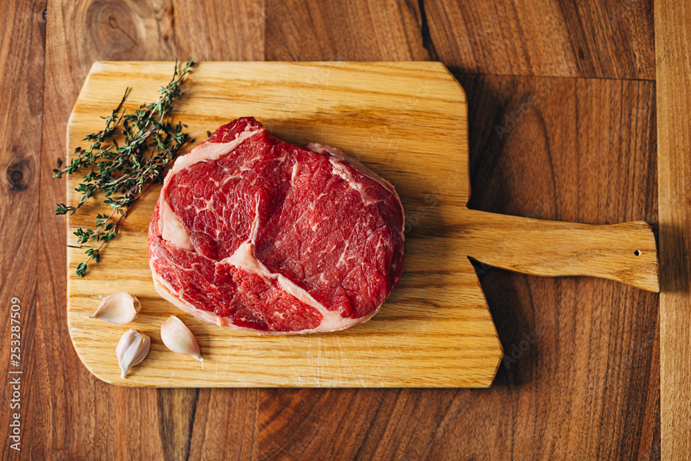 Overhead shot of ribeye steak resting on a wooden board in the kitchen with fresh thyme and garlic cloves. Top shot Overhead shot of ribeye steak resting on a wooden board in the kitchen with fresh thyme and garlic cloves. Top shot