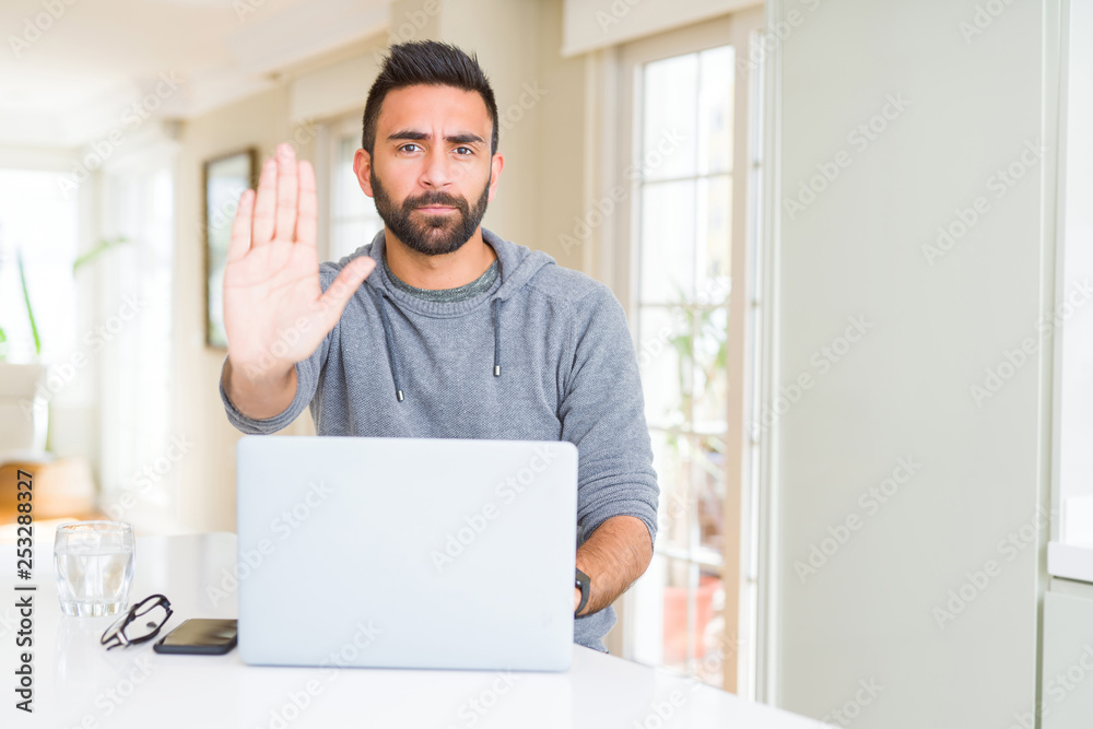 Handsome hispanic man working using computer laptop with open hand doing stop sign with serious and confident expression, defense gesture Handsome hispanic man working using computer laptop with open hand doing stop sign with serious and confident expression, defense gesture