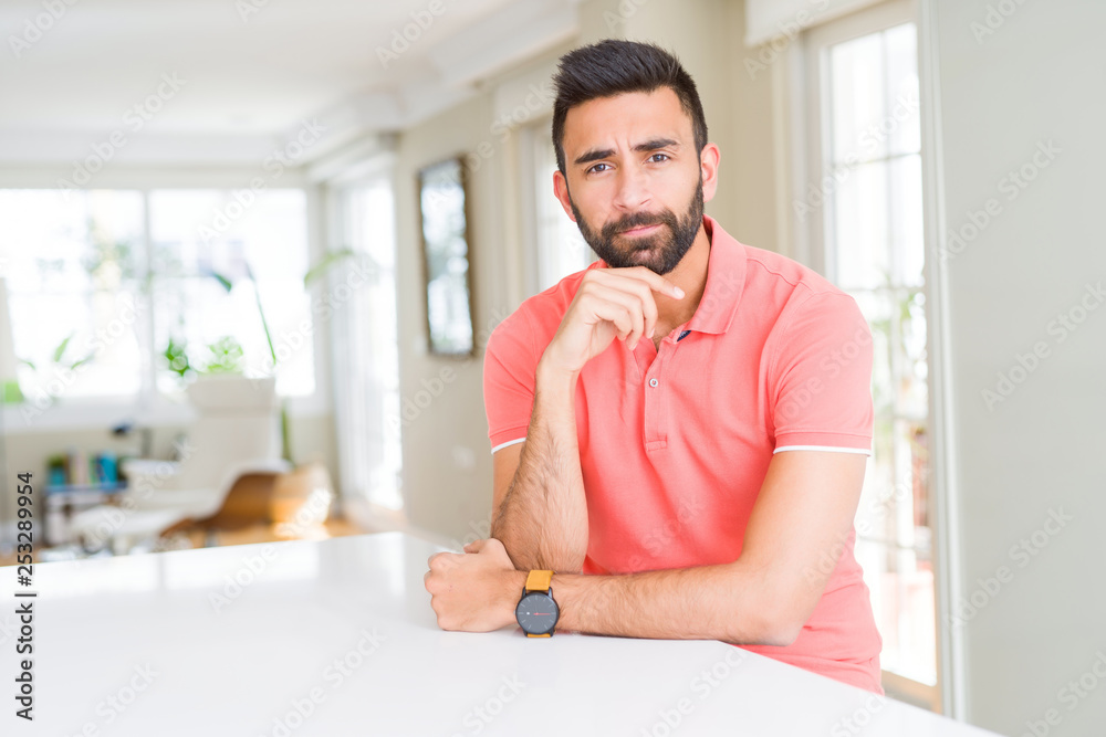 Handsome hispanic man wearing casual t-shirt at home with hand on chin thinking about question, pensive expression. Smiling with thoughtful face. Doubt concept.