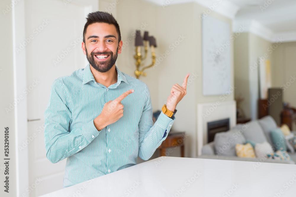 Handsome hispanic man at home smiling and looking at the camera pointing with two hands and fingers to the side.