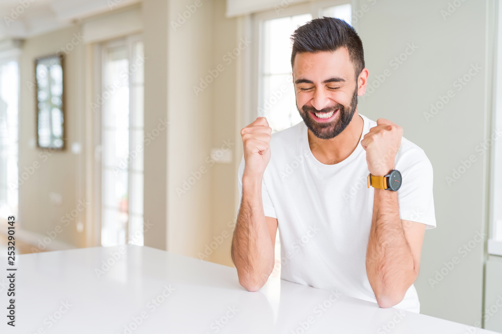 Handsome hispanic man casual white t-shirt at home excited for success with arms raised celebrating victory smiling. Winner concept. Handsome hispanic man casual white t-shirt at home excited for success with arms raised celebrating victory smiling. Winner concept.