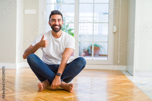 Handsome hispanic man wearing casual t-shirt sitting on the floor at home doing happy thumbs up gesture with hand. Approving expression looking at the camera with showing success.