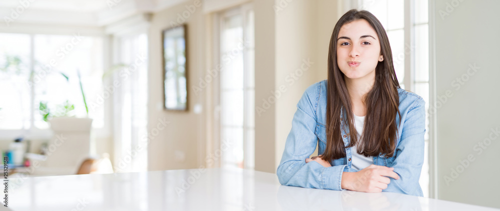 Wide angle picture of beautiful young woman sitting on white table at home puffing cheeks with funny face. Mouth inflated with air, crazy expression. Wide angle picture of beautiful young woman sitting on white table at home puffing cheeks with funny face. Mouth inflated with air, crazy expression.