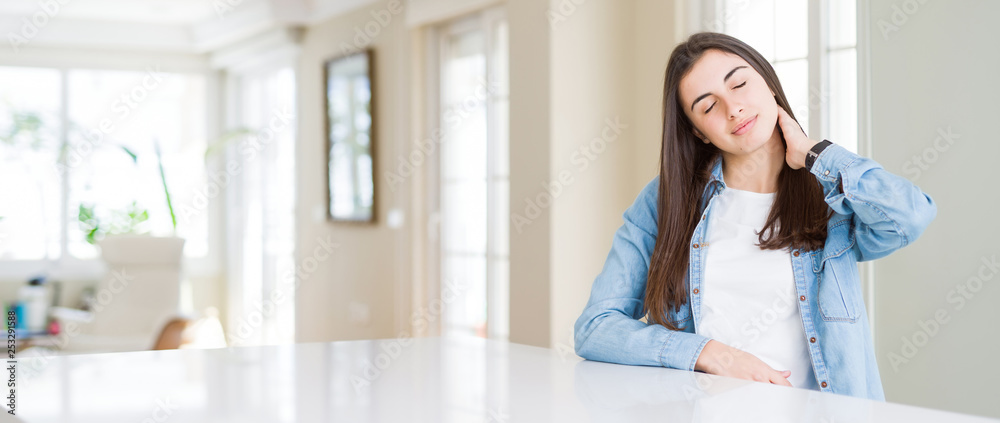 Wide angle picture of beautiful young woman sitting on white table at home Suffering of neck ache injury, touching neck with hand, muscular pain