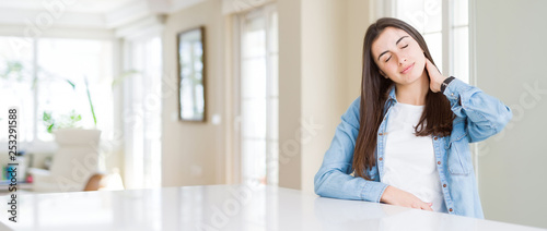 Wide angle picture of beautiful young woman sitting on white table at home Suffering of neck ache injury  touching neck with hand  muscular pain