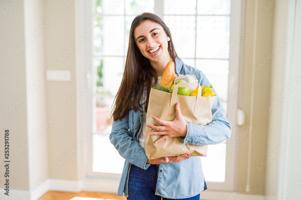 Beautiful young woman holding paper bag full of healthy groceries with a happy face standing and smiling with a confident smile showing teeth Beautiful young woman holding paper bag full of healthy groceries with a happy face standing and smiling with a confident smile showing teeth