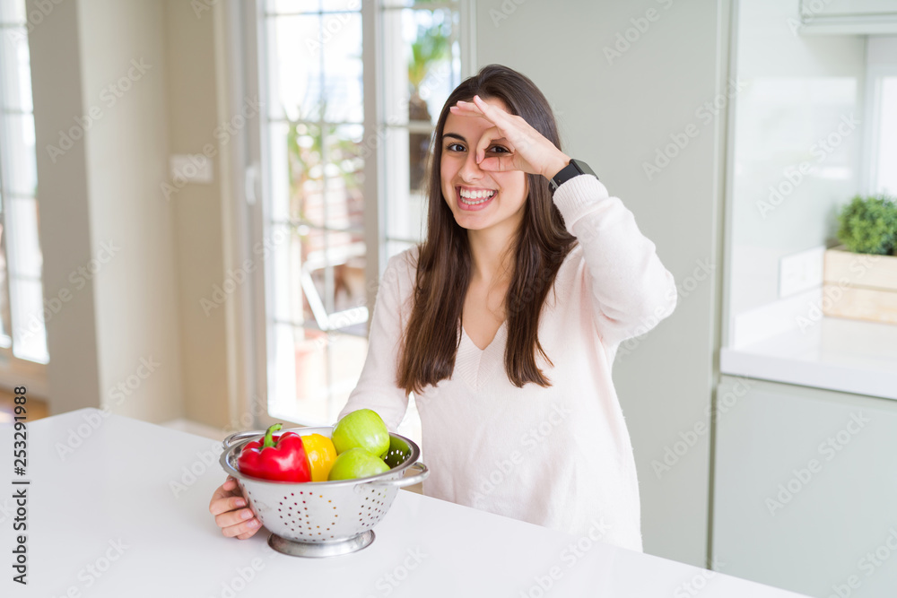 Beautiful young woman using colander to wash and clean vegatables with happy face smiling doing ok sign with hand on eye looking through fingers