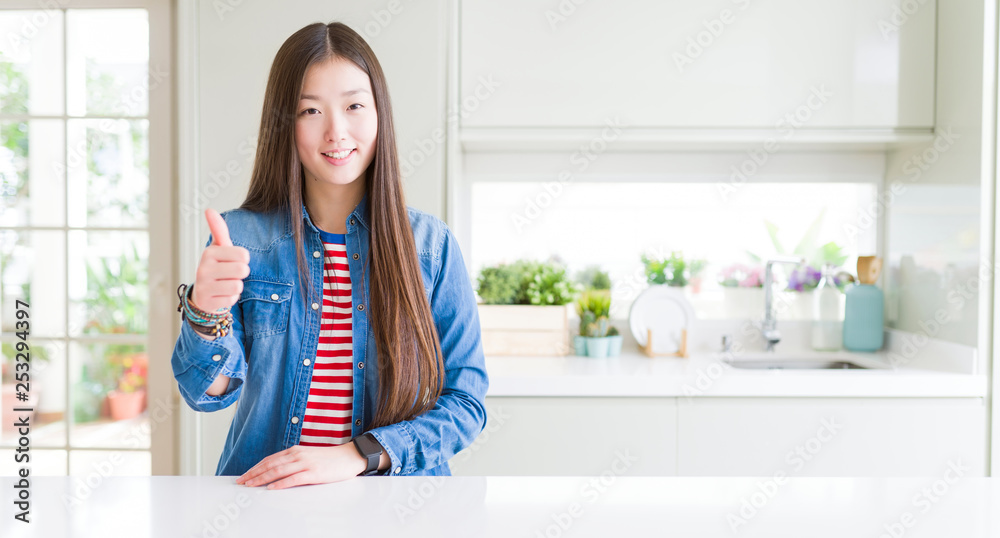 Beautiful Asian woman wearing denim jacket on white table doing happy thumbs up gesture with hand. Approving expression looking at the camera showing success. Beautiful Asian woman wearing denim jacket on white table doing happy thumbs up gesture with hand. Approving expression looking at the camera showing success.