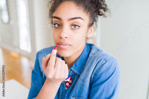 Young african american woman applying red lipstick on lips with a confident expression on smart face thinking serious