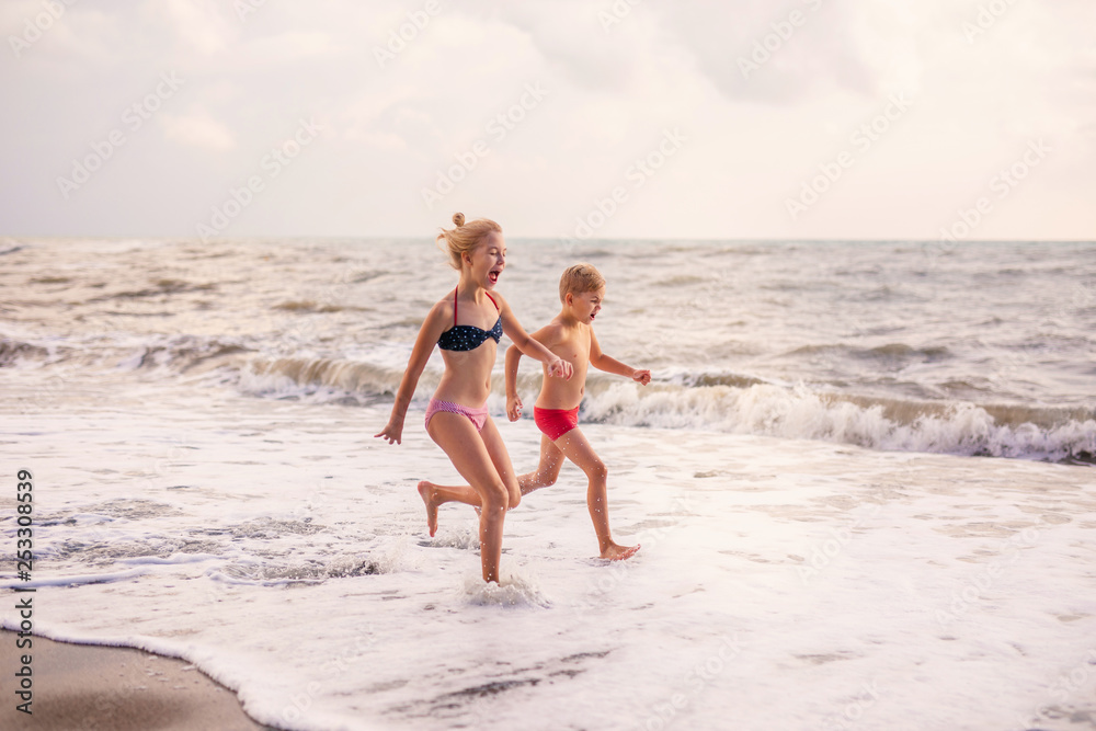 Cute blonde children boy and girl running from sea waves on beach