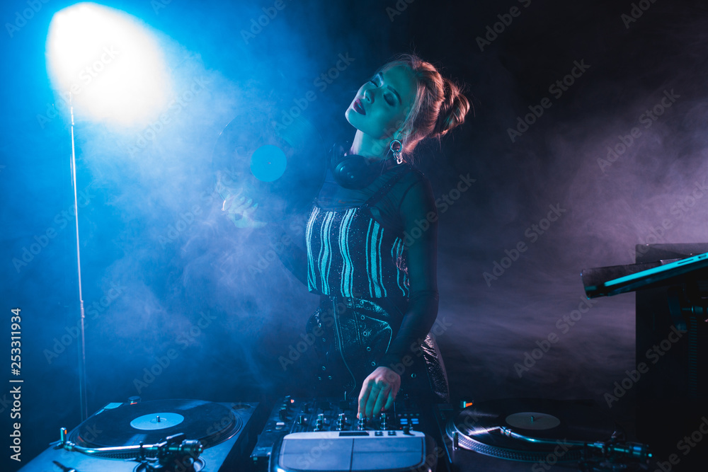 blonde dj woman standing with closed eyes near dj equipment and holding  retro vinyl record in nightclub with smoke Stock Photo | Adobe Stock