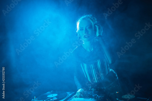 attractive dj girl listening music in headphones near dj equipment in nightclub with smoke