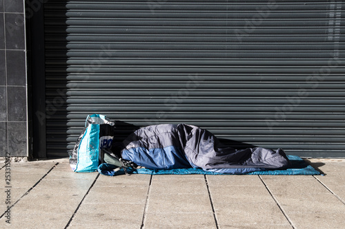 Homeless person asleep in a sleeping bag on a pavement sidewalk in front of a metal shutter.  Face not visable.