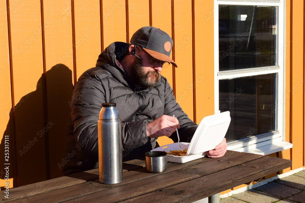 A man is eating lunch at a roadside cafe. A bearded man with glasses eats french fries and drinks coffee from a thermos. Cafe by the road. Norway. A man is eating lunch at a roadside cafe. A bearded man with glasses eats french fries and drinks coffee from a thermos. Cafe by the road. Norway.