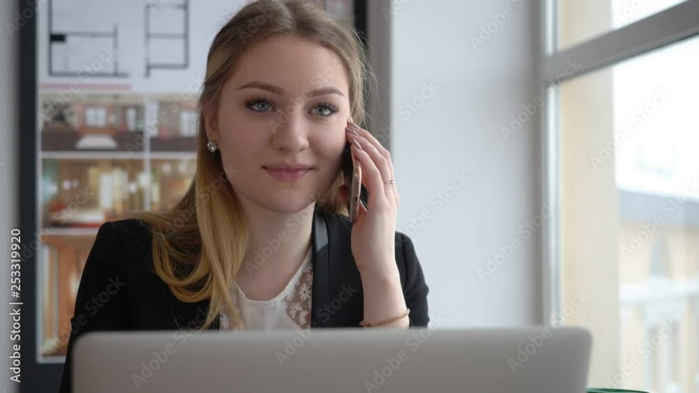 Young woman manager talking by mobile phone working on laptop in business office. Creative designer talking to smartphone with client in business office. Business woman using mobile for conversation Young woman manager talking by mobile phone working on laptop in business office. Creative designer talking to smartphone with client in business office. Business woman using mobile for conversation