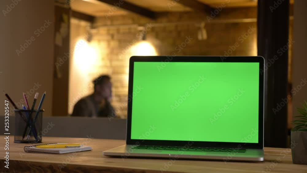Closeup shoot of a green chroma screen of the laptop lying on the desk in the office indoors with an employee typing on the blurred background Closeup shoot of a green chroma screen of the laptop lying on the desk in the office indoors with an employee typing on the blurred background