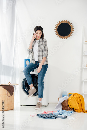woman sitting on washer and talking on smartphone in laundry room