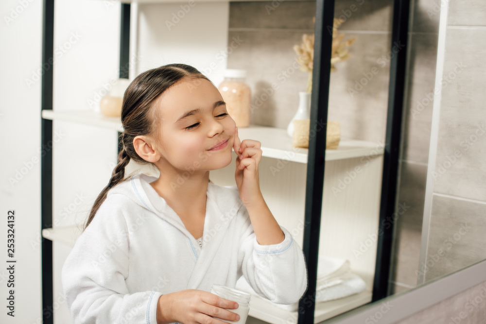 selective focus child in white bathrobe looking to mirror and applying cosmetic cream in bathroom selective focus child in white bathrobe looking to mirror and applying cosmetic cream in bathroom