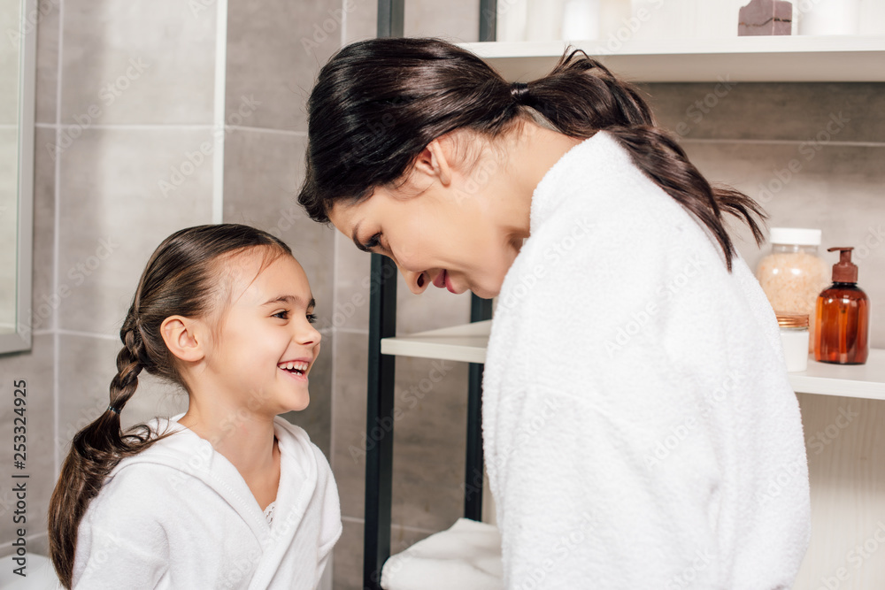 mother looking to smiling daughter in white bathrobe near shelves in bathroom mother looking to smiling daughter in white bathrobe near shelves in bathroom