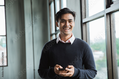 asian young man smiling in the office room look at camera with hold a smartphone