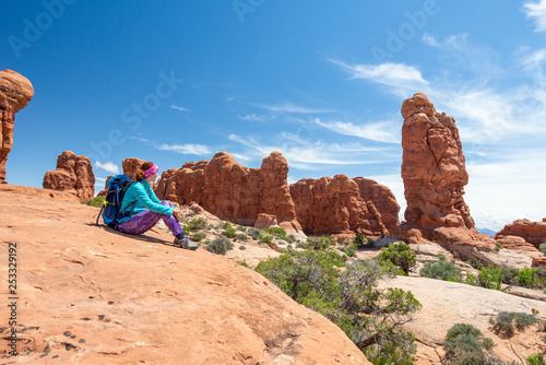 Young woman wearing backpack is sitting and looking at the Garden of Eden in Arches National Park in Utah. Female hiker in Arches National Park. Travel and adventure concept.
