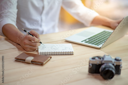 Male illustrator hand drawing world map outline on notebook paper working with laptop computer and film camera on wooden table. Graphic designer creating artwork for online travel content. photo