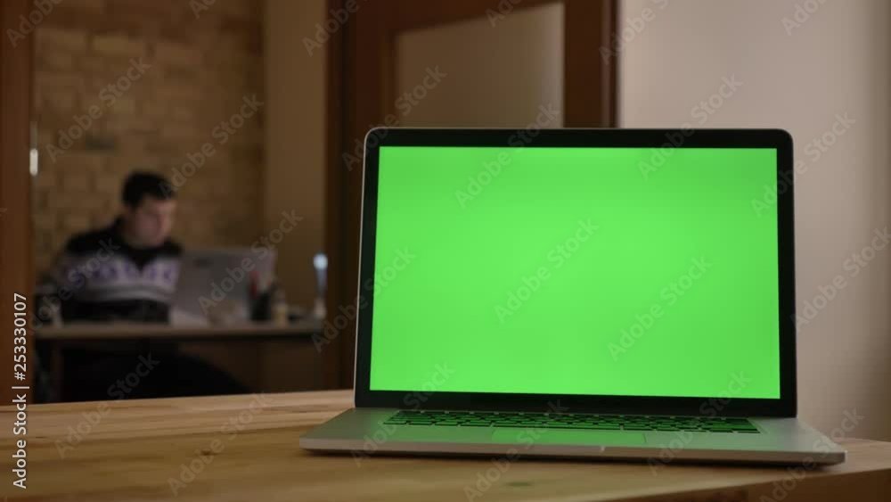 Closeup shoot of green screen of the laptop laying on the desk in office indoors with businessman working on the blurred background Closeup shoot of green screen of the laptop laying on the desk in office indoors with businessman working on the blurred background
