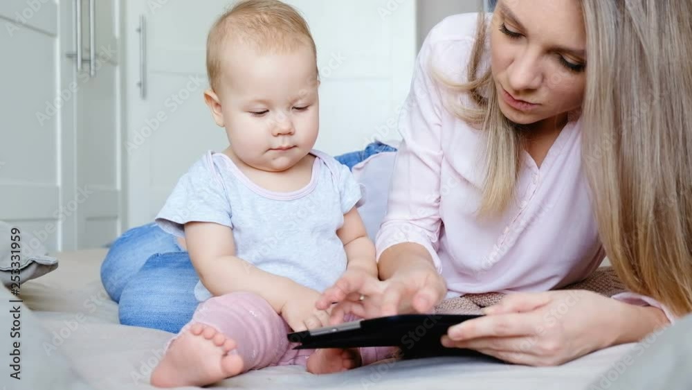 Young cute mom shows her child educational games on a tablet during a weekend relaxing in the bedroom in a comfortable home environment. Concept of educational games for kids Young cute mom shows her child educational games on a tablet during a weekend relaxing in the bedroom in a comfortable home environment. Concept of educational games for kids