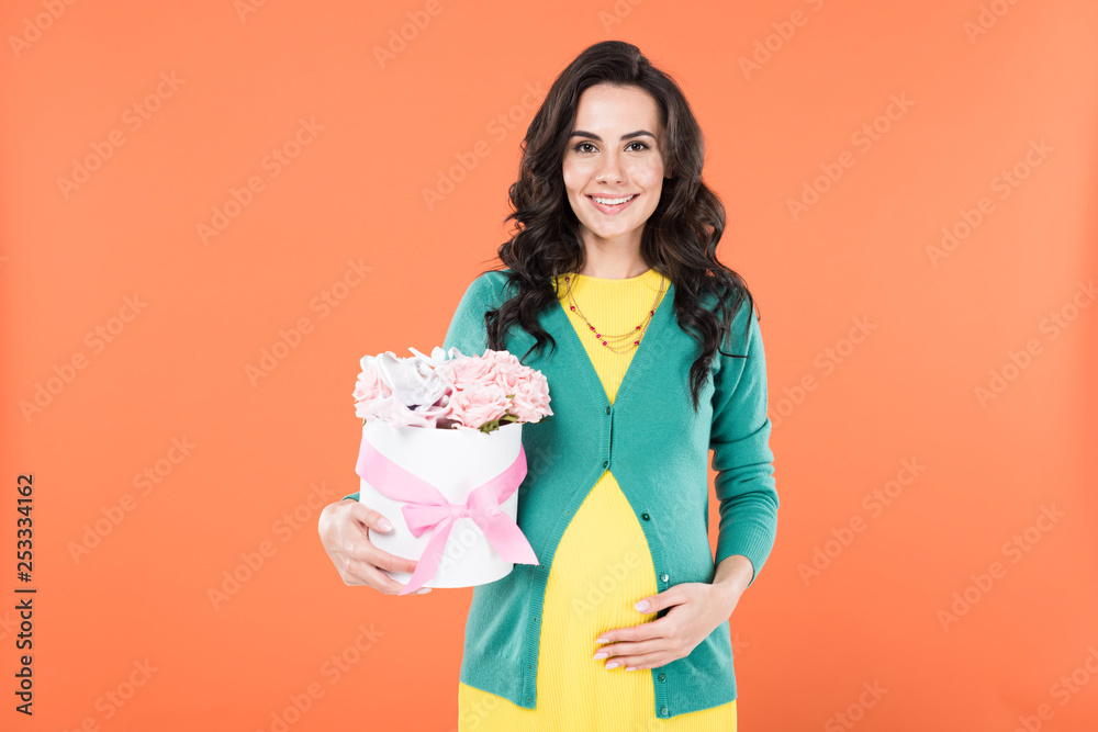 Attractive pregnant woman holding pink flowers and touching belly isolated on orange Attractive pregnant woman holding pink flowers and touching belly isolated on orange