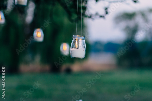 a burning candle in a closed candlestick made of glass hangs outside in a park
