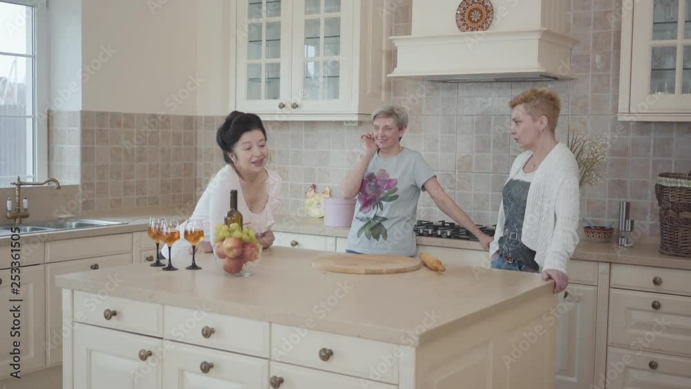 Senior ladies having fun spending time together drinking wine and eating fruits. Three mature women communicating chatting at home standing near modern table in the middle of kitchen. Senior ladies having fun spending time together drinking wine and eating fruits. Three mature women communicating chatting at home standing near modern table in the middle of kitchen.
