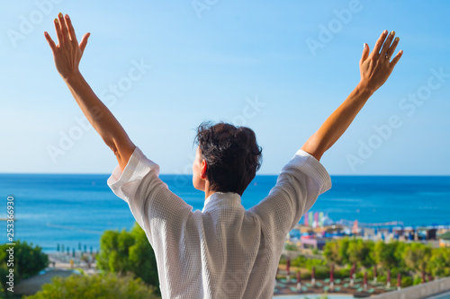 Relaxed young woman in white cozy bathrobe standing on balcony in summer morning. brunette girl on summer vacation.Woman in bath robe looking at beautiful ocean view from a tropical balcony,patio