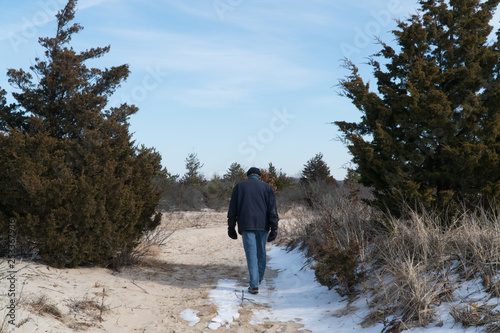 Elderly man walking alone through trail in the woods past tree gap on a cold winter day after snow fall on path. Day time exterior