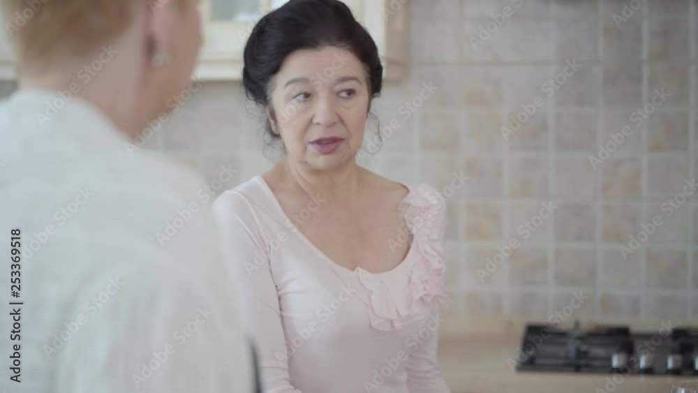Middle aged mature women communicating chatting in the kitchen at home. Lady telling story to her friends emotionally, actively gesticulating. Middle aged mature women communicating chatting in the kitchen at home. Lady telling story to her friends emotionally, actively gesticulating.