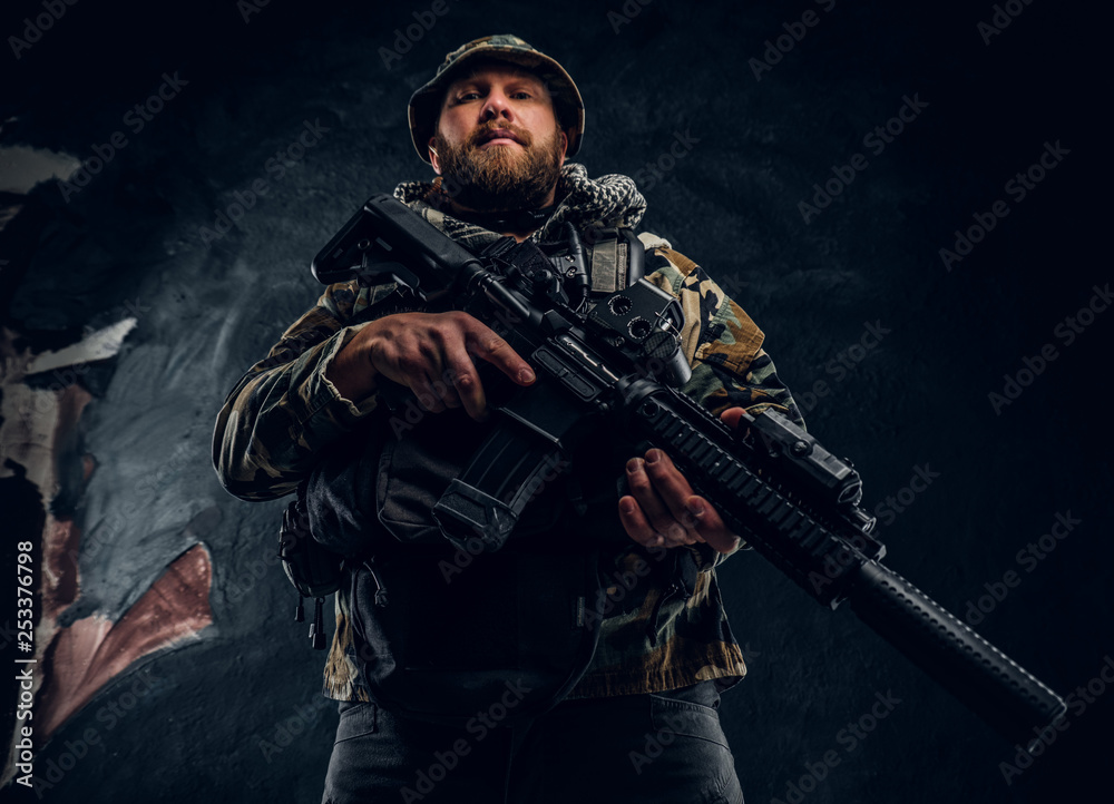 Brutal man in the military camouflaged uniform holding an assault rifle. Studio photo against a dark textured wall Brutal man in the military camouflaged uniform holding an assault rifle. Studio photo against a dark textured wall