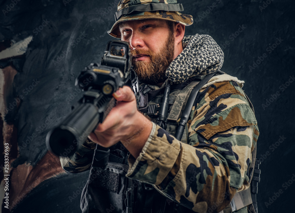 Brutal man in the military camouflaged uniform holding an assault rifle with a laser sight and aims at the target. Studio photo against a dark textured wall