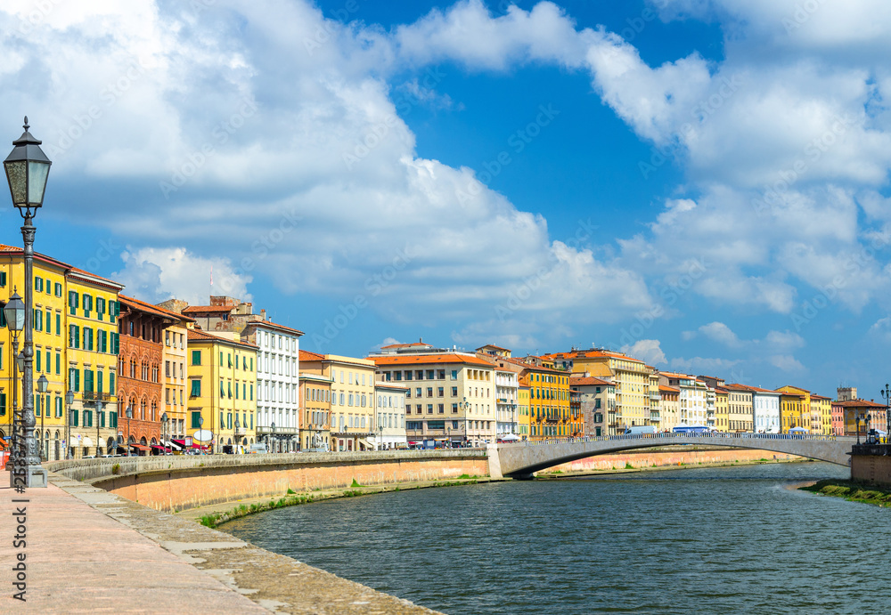 Row of old colorful buildings houses on embankment promenade of Arno river, Ponte Di Mezzo bridge in historical centre of Pisa town with blue sky white clouds background in sunny day, Tuscany, Italy