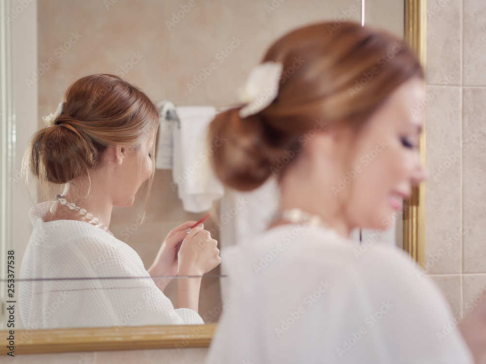 Beautiful European woman in a white dressing gown is standing near the mirror and using nail file. Beautiful European woman in a white dressing gown is standing near the mirror and using nail file.