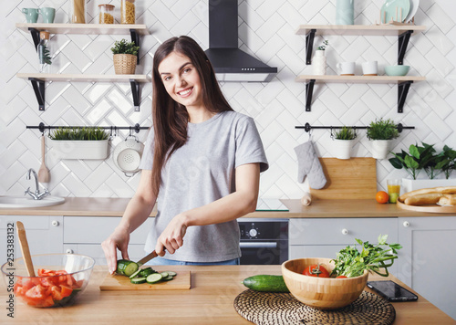 Happy young girl prepares healthy food in the kitchen slicing cucumber