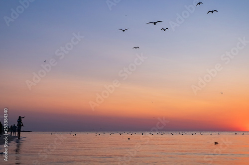 people meet the colorful sunrise on the beach to the sea. silhouettes of people and seagulls. father holds a child on his shoulders © AlexLit