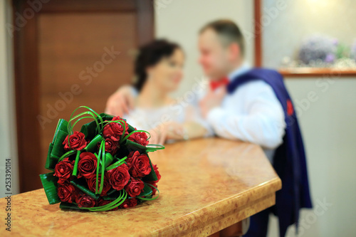 Bukiet ślubny z czerwonych róż i młoda para w tle.