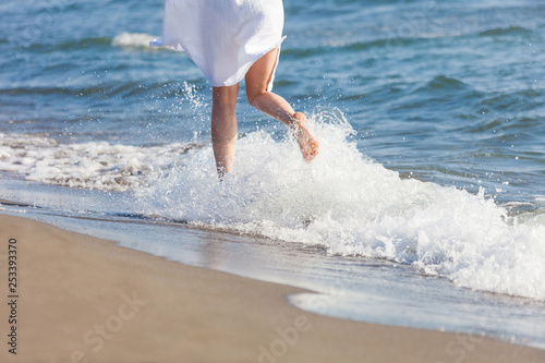 woman in white cotton dress run through water on sandy sea beach sunny summer day lower body back view
