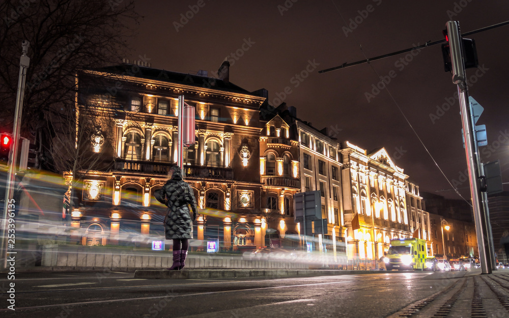 Fototapeta premium Long Exposure Shot of a Woman Crossing a Busy Street in Dublin Ireland - with Cars Driving Past in the Background