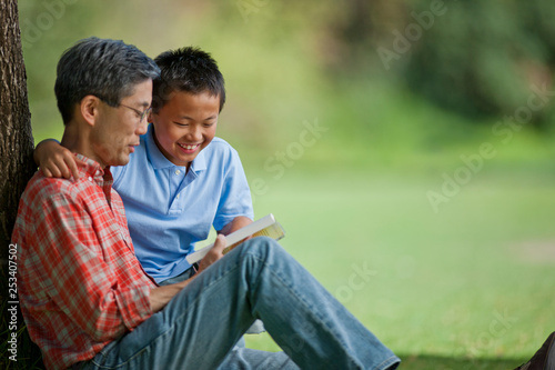 Happy father and son reading a book under a tree.